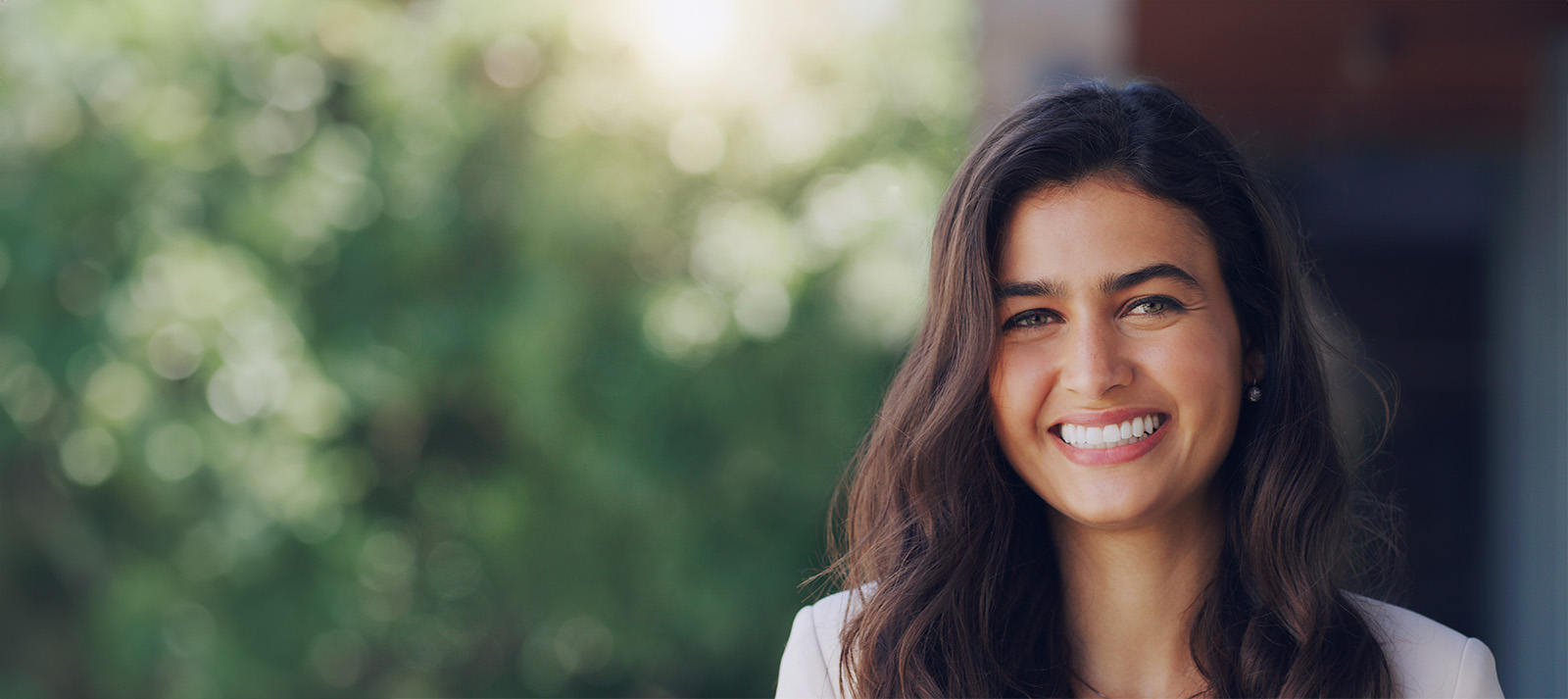A woman with long hair smiles at the camera, set against a blurred background featuring greenery and a building.