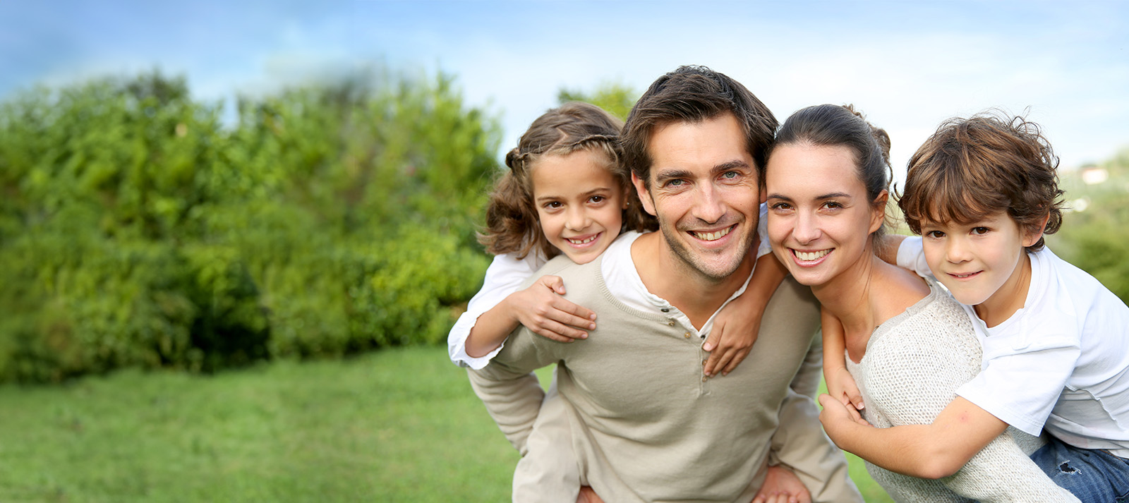 The image shows a family of four people outdoors during daylight, with the father standing in front of his wife and two children, all smiling and looking towards the camera.